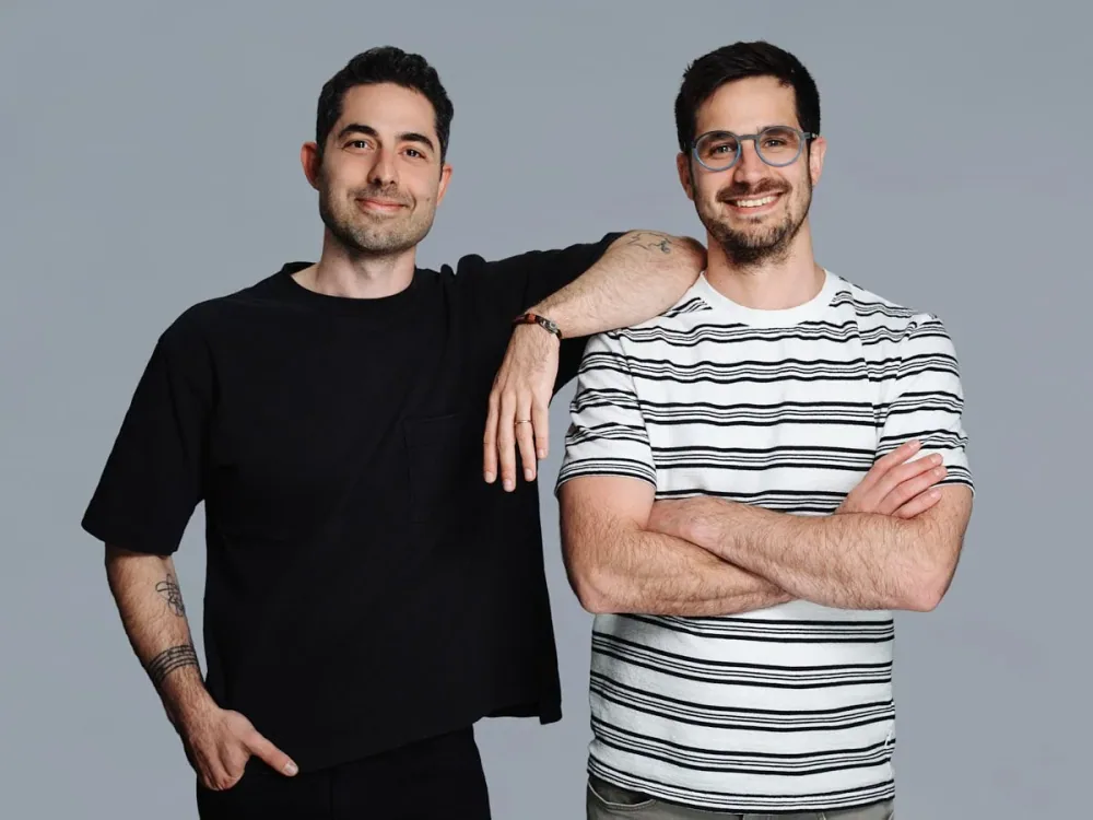 Two men standing together against a grey background.
