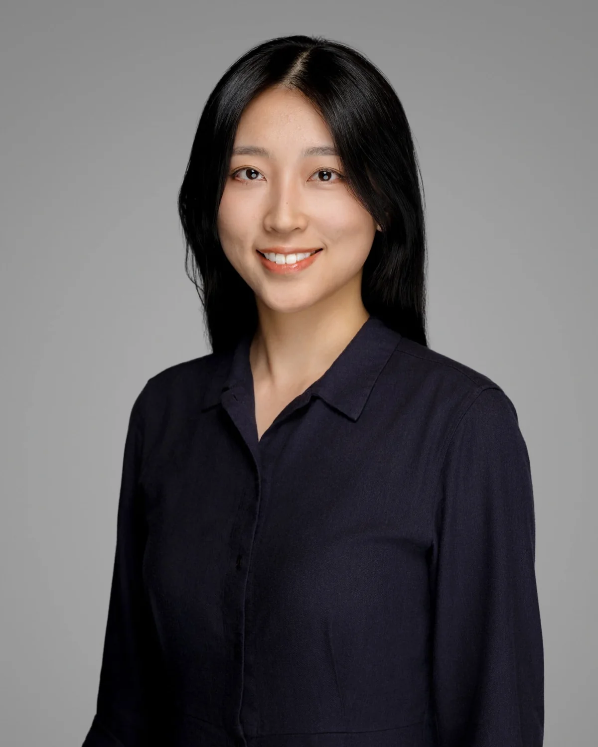 A person in a dark buttoned shirt, centered against a gray backdrop.