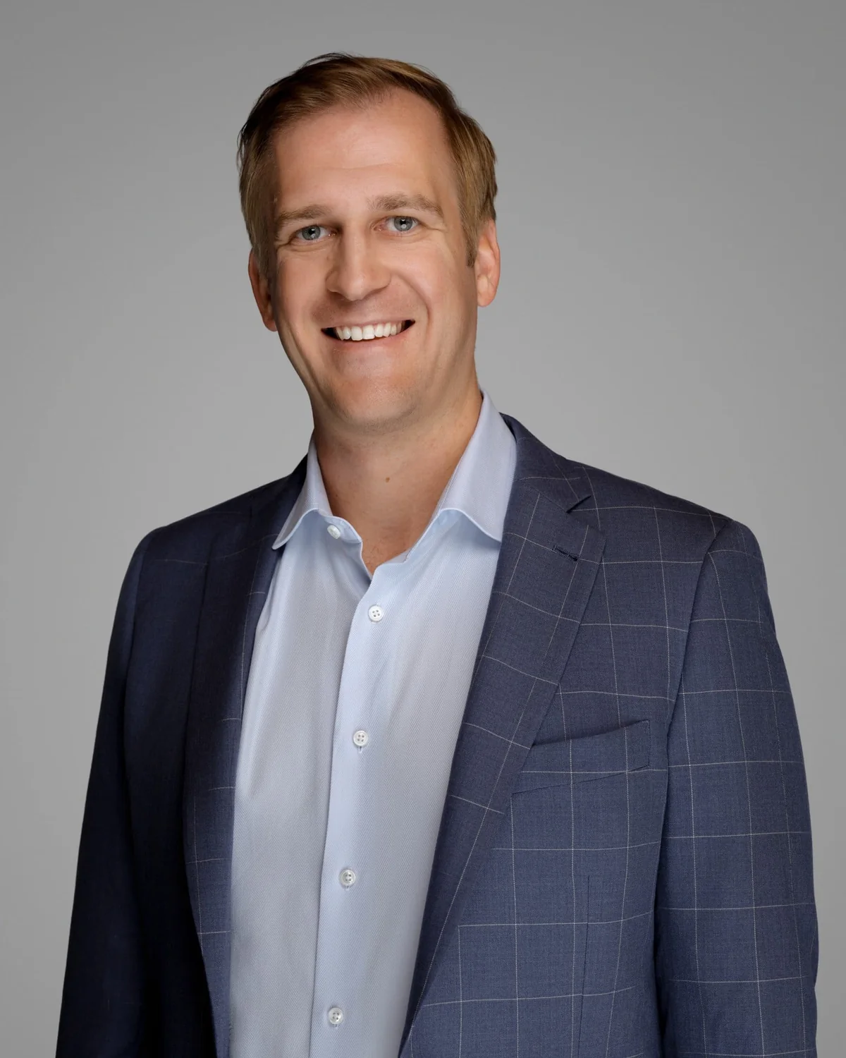 A person in a blue checked jacket over a light shirt, standing against a gray backdrop.