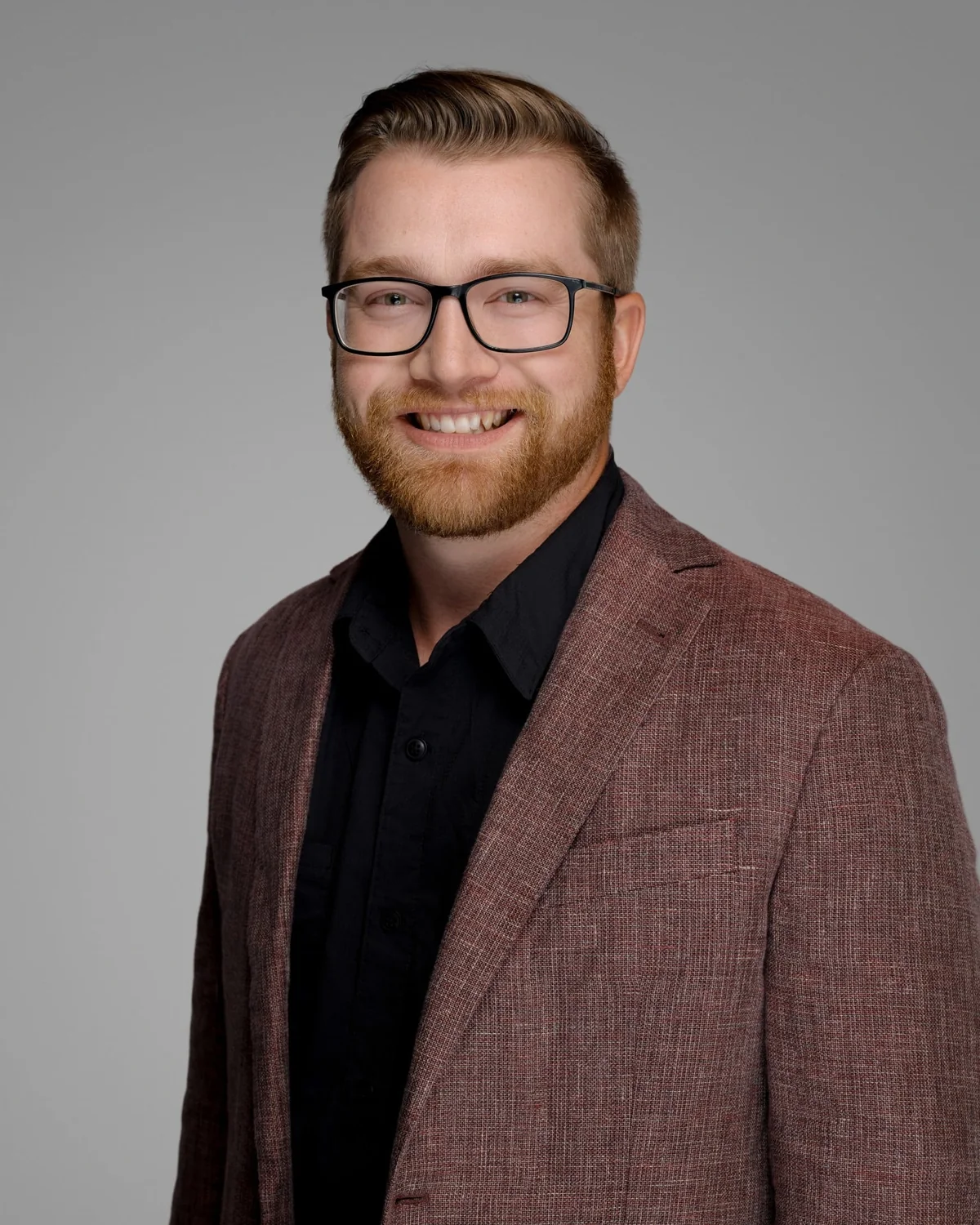 A person wearing glasses, a patterned burgundy jacket and a black shirt against a gray studio backdrop.