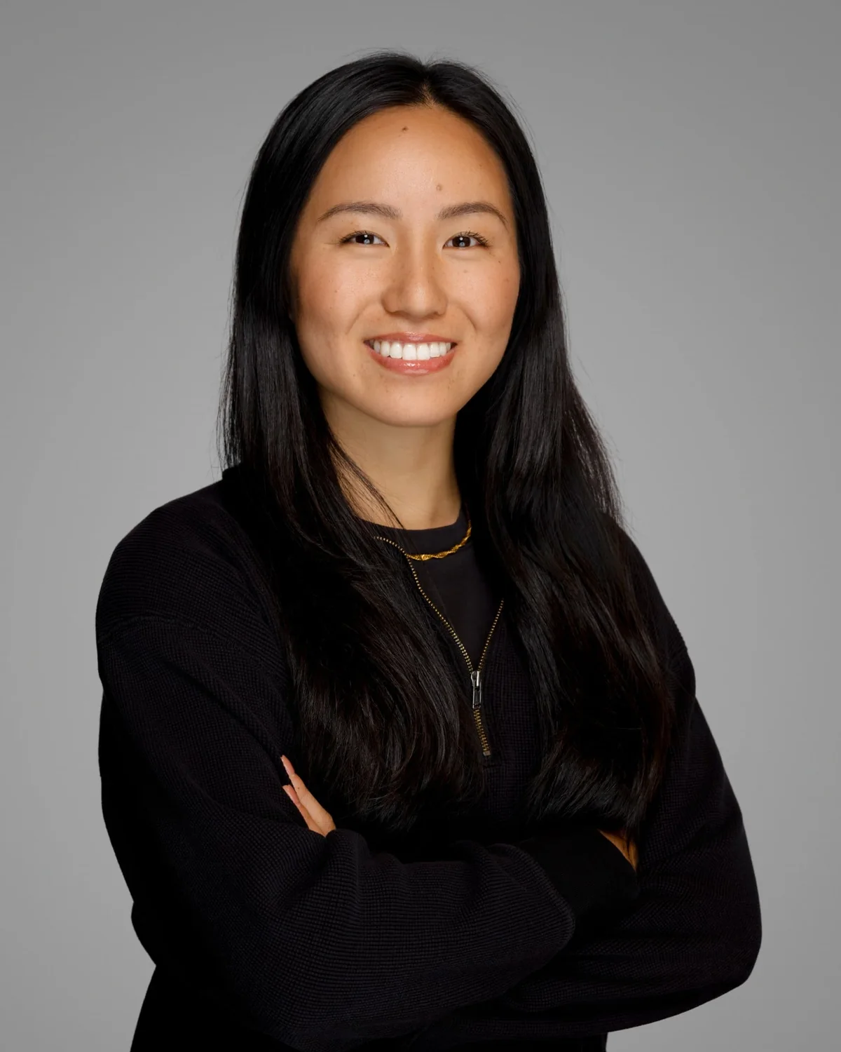 A person in a black long-sleeve top, standing with arms folded against a gray studio backdrop.