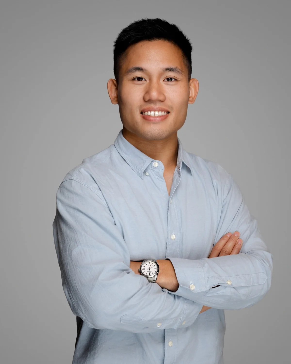 A person in a light blue buttoned shirt, standing with arms folded against a gray studio backdrop.